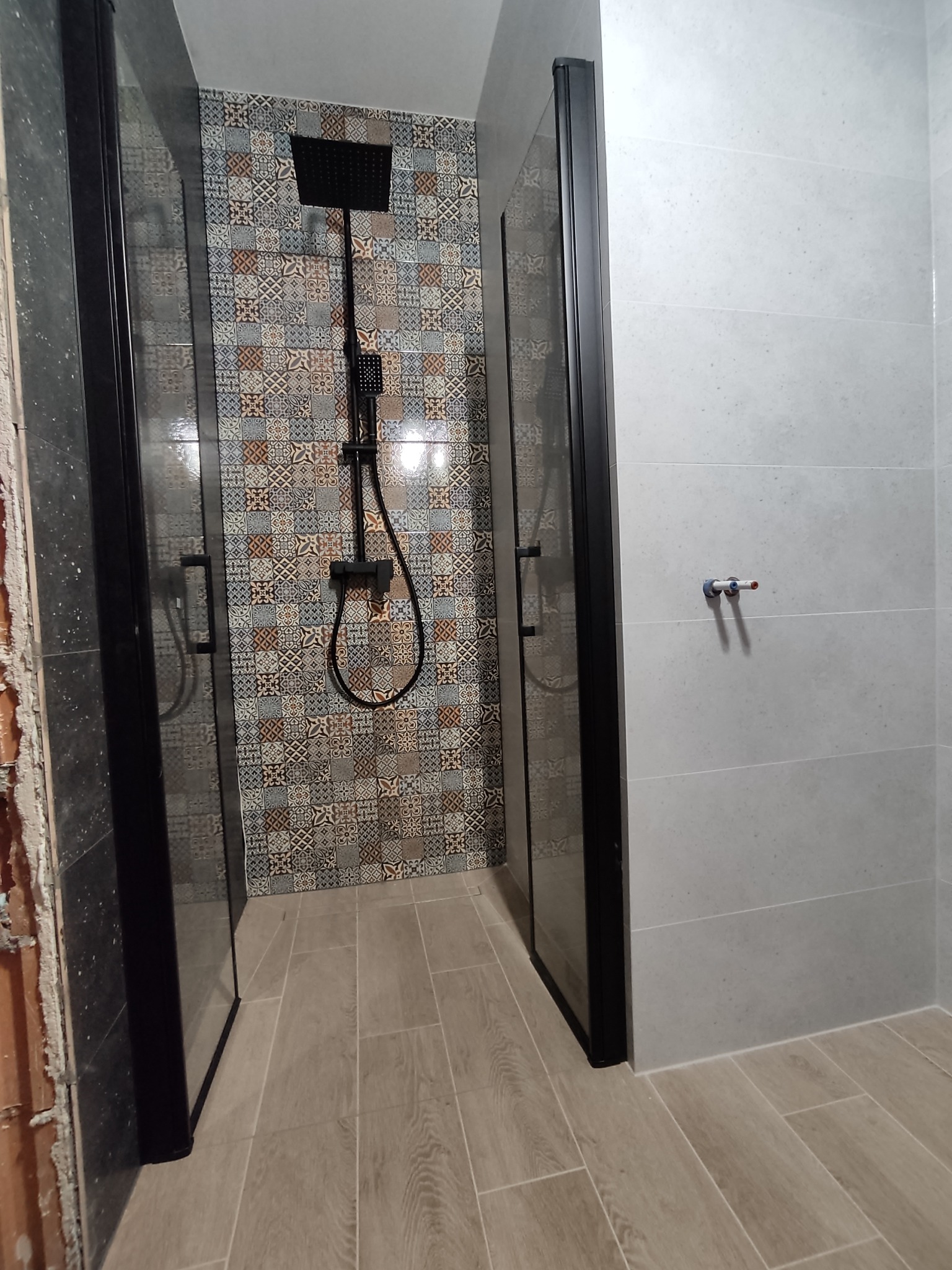 Nowoczesna kabina prysznicowa typu walk-in z czarnymi profilami, wykończona płytkami w dwóch stylach: patchworkowym pod prysznicem i gładkimi szarymi na pozostałych ścianach.