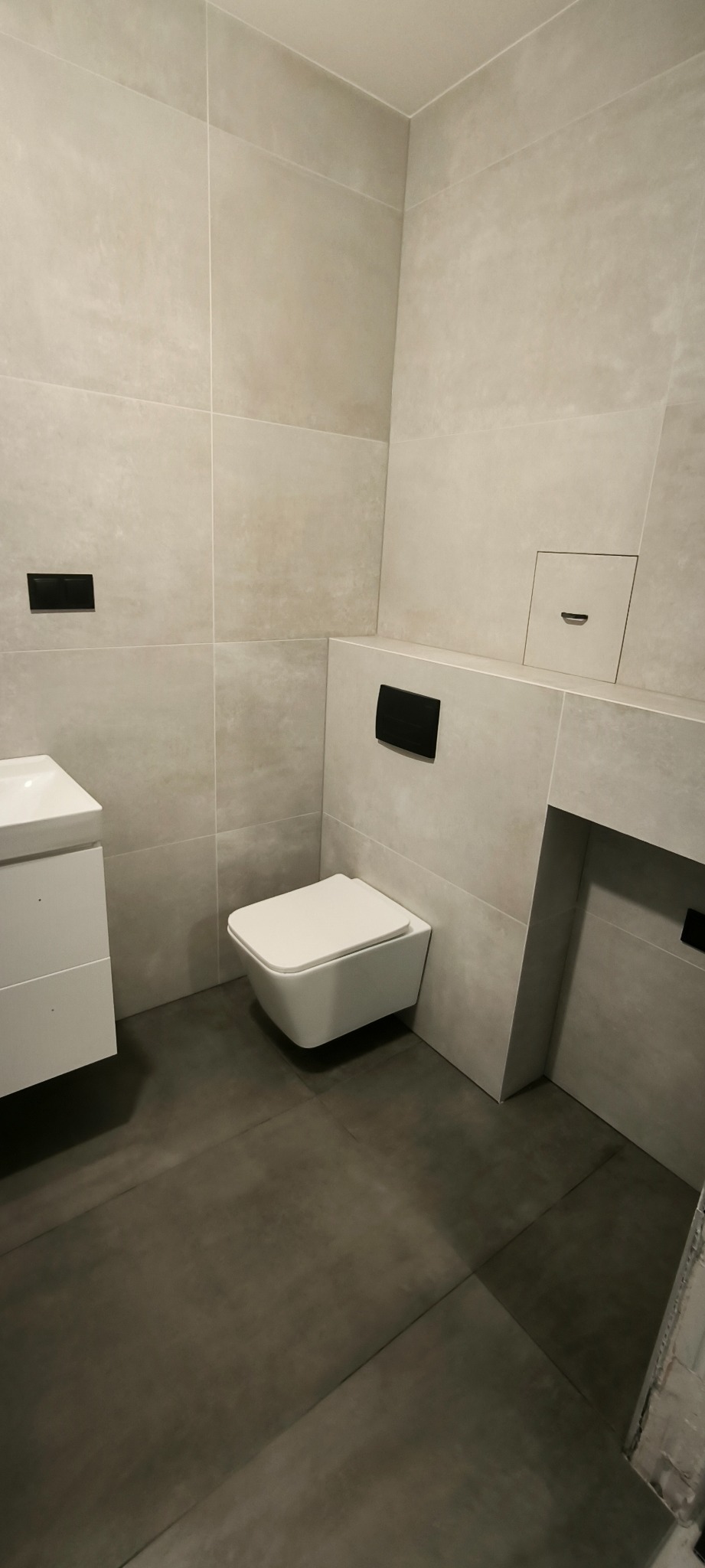 Minimalistyczna łazienka z wiszącą toaletą, umywalką i szarymi płytkami imitującymi beton, podkreślona czarnymi akcentami w postaci przycisków spłukiwania.
