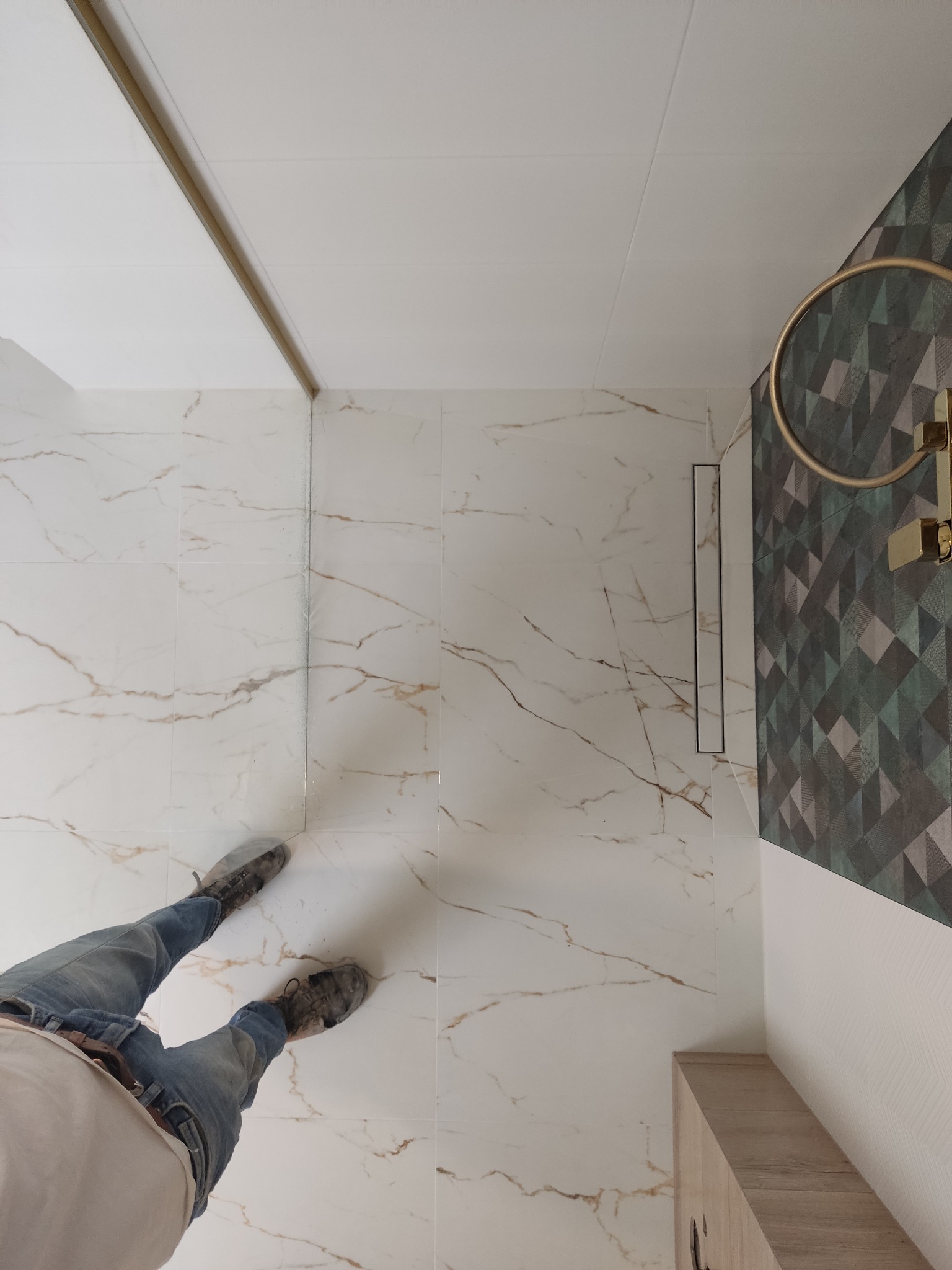 Widok z góry na wykończoną łazienkę z białymi płytkami imitującymi marmur, odpływem liniowym i fragmentem osoby w jeansach i roboczych butach.