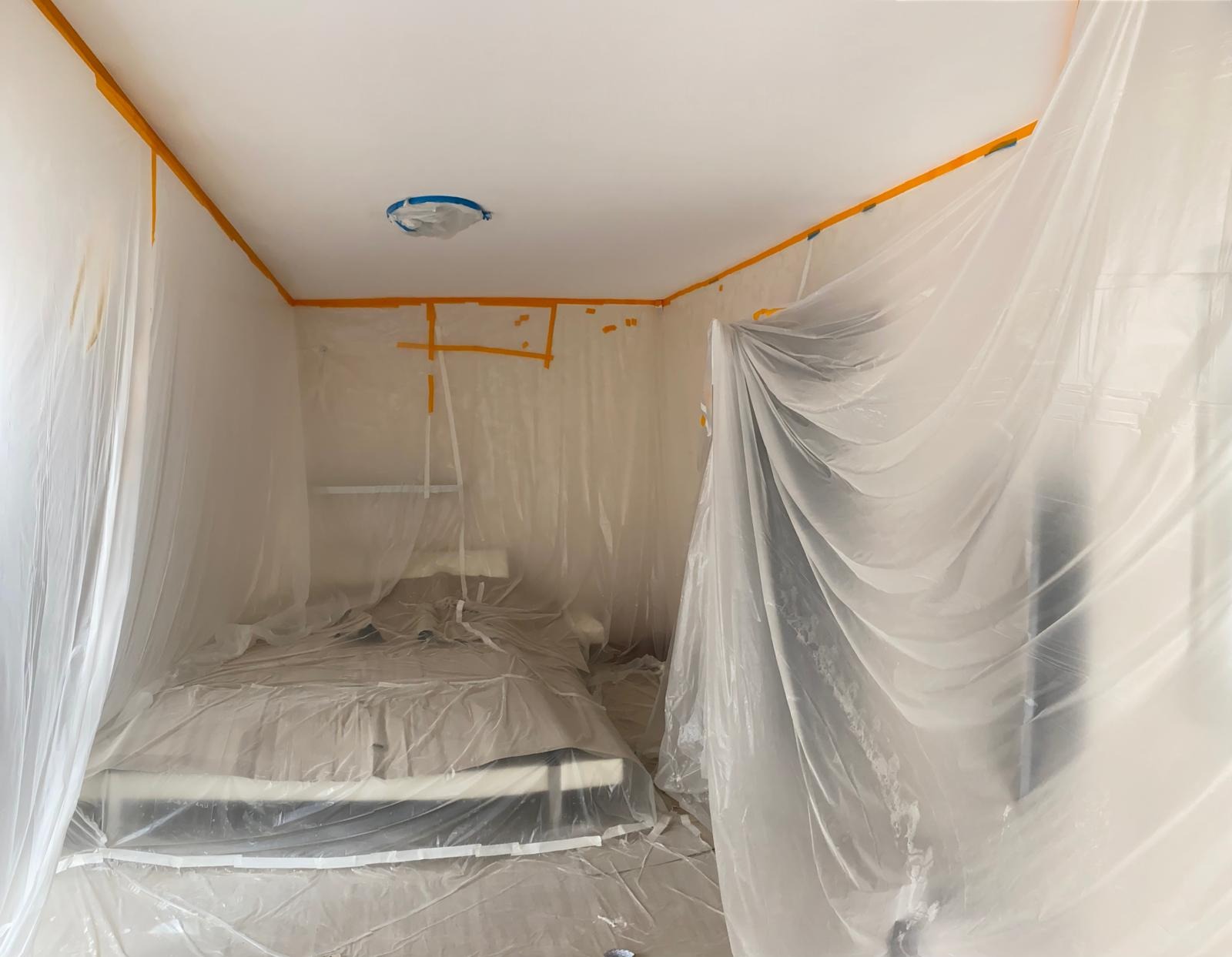 Pokój zabezpieczony folią malarską i taśmą przed pracami wykończeniowymi, widoczny zakryty mebel i lampa na suficie.