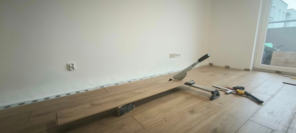 Przycinanie panelu laminowanego za pomocą gilotyny do paneli, widoczne listwy przypodłogowe, poziomica, miarka i młotek na podłodze w trakcie montażu.