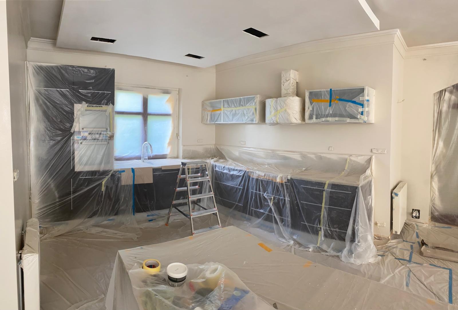 Kuchnia w trakcie zabezpieczania folią malarską przed pracami wykończeniowymi, widoczne zakryte meble kuchenne, okno, grzejnik, drabina oraz pojemniki z farbą i taśma malarska.