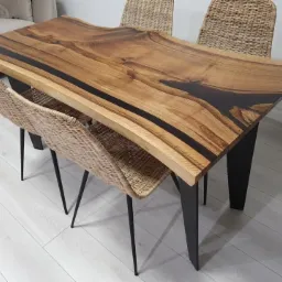 Stół z blatem z litego drewna z żywicą epoksydową w kolorze ciemnego brązu, na czarnych, metalowych nogach, otoczony plecionymi krzesłami na cienkich, czarnych nogach, ustawiony na jasnej podłodze.