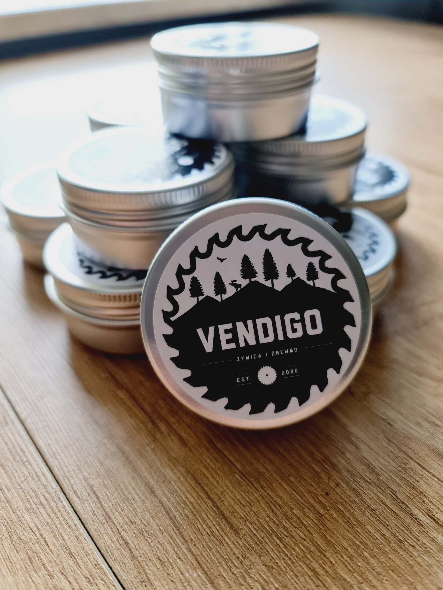 Stos metalowych puszek z etykietą 'Vendigo Żywica i Drewno' na drewnianym blacie, logo z motywem piły, drzew i jelenia.