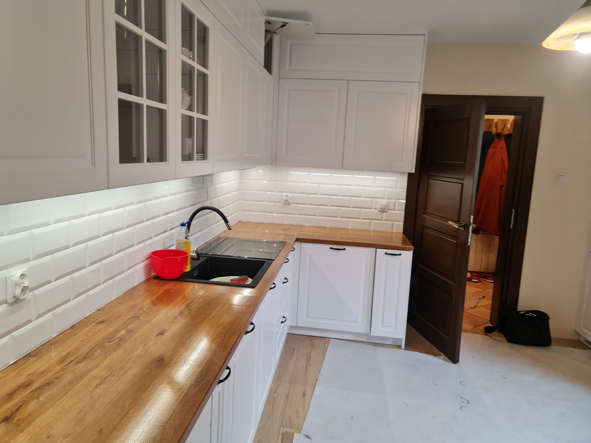 Nowoczesna kuchnia z białymi szafkami i drewnianym blatem, widok na otwarte drzwi do innego pomieszczenia z wiszącymi ubraniami.