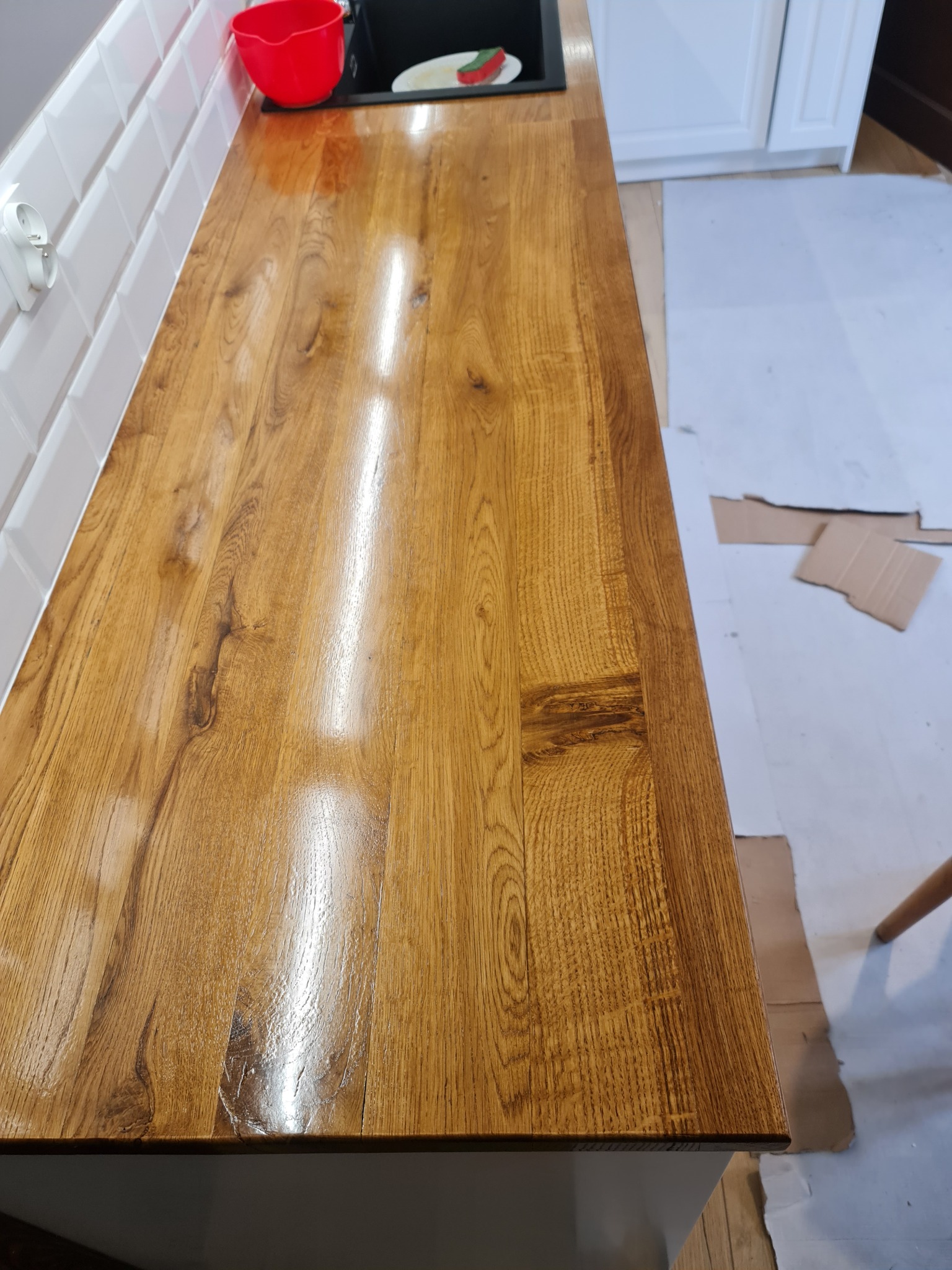 Świeżo zamontowany, lakierowany blat drewniany o ciepłym odcieniu, widoczne słoje drewna i odbicia światła, białe płytki na ścianie w tle, fragment zlewu i czerwona miska, zabezpieczenie podłogi...