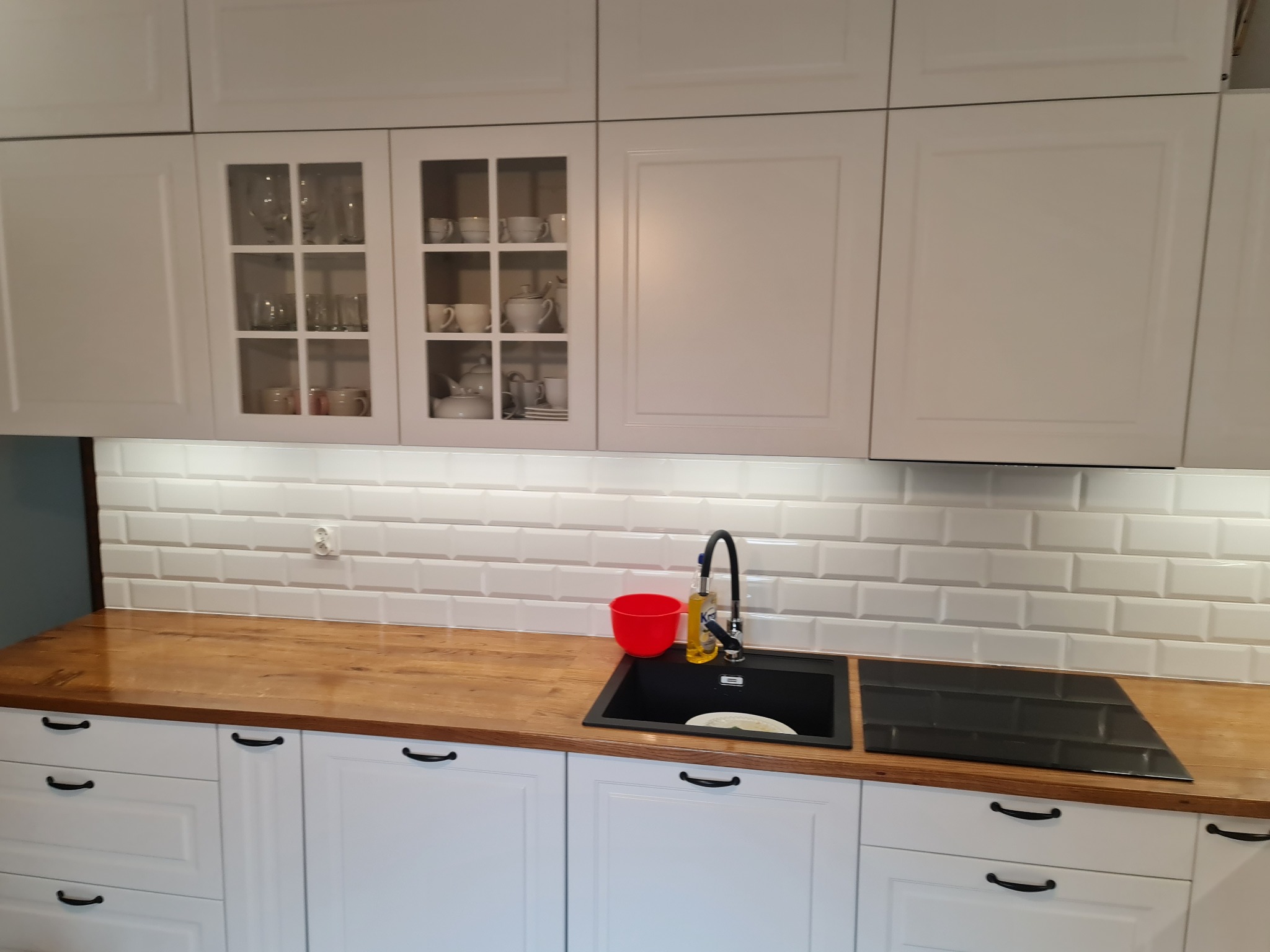 Kuchnia z białymi szafkami górnymi, niektóre z przeszkleniami, blatem z drewna, czarnym zlewem i płytą indukcyjną, oraz białymi płytkami na ścianie.