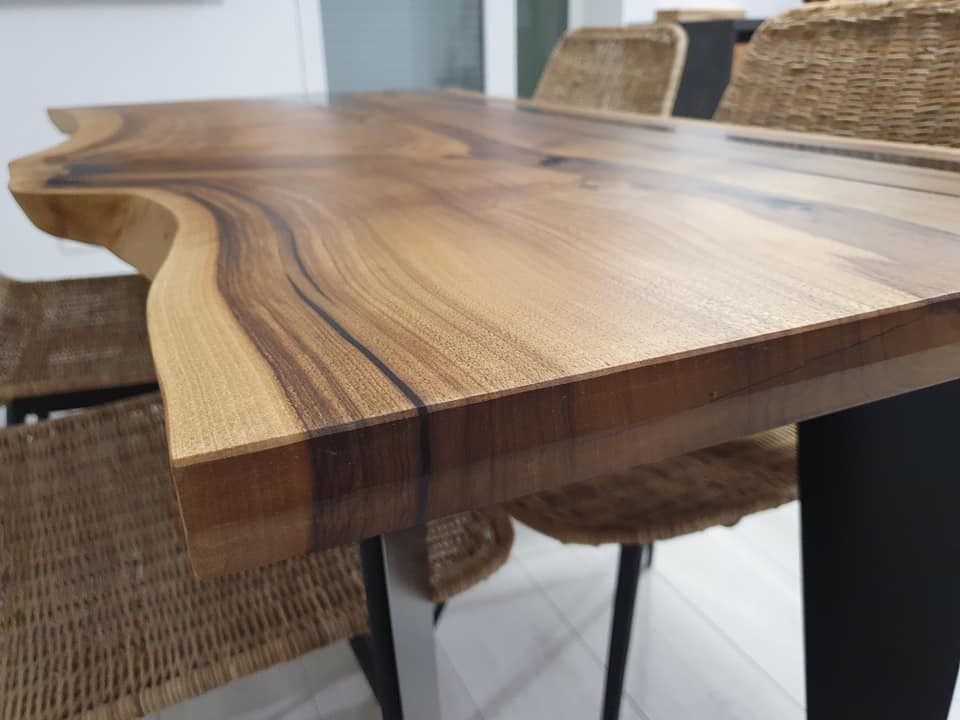 Blat stołu z litego drewna o nieregularnym kształcie krawędzi, wykończony lakierem, wsparty na czarnych metalowych nogach, w tle wiklinowe krzesła.