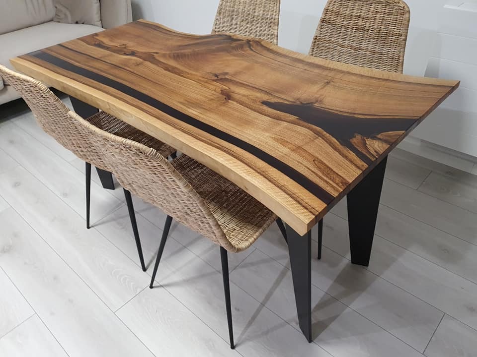 Stół z blatem z litego drewna z żywicą epoksydową w kolorze ciemnego brązu, na czarnych, metalowych nogach, otoczony plecionymi krzesłami na cienkich, czarnych nogach, ustawiony na jasnej podłodze.