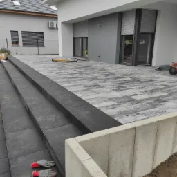 Wykończony taras z szaro-białej kostki brukowej z trzema stopniami, obok domu z szarymi roletami, widoczne narzędzia budowlane i rękawice robocze.