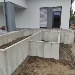 Fundamenty pod budynek z szarych bloczków betonowych, widoczne wykopane rowy wypełnione ziemią, na pierwszym planie błotniste podłoże.