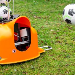 WYTRENUJ Michał Sadyk - Pomarańczowa maszyna do wyrzucania piłek na trawie, z piłkami na górze i jedną w locie, widoczny napis 'ball-launcher.com'.