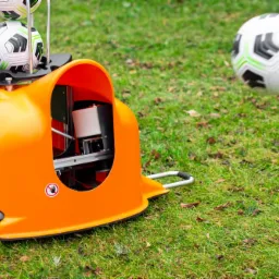 Pomarańczowa maszyna do wyrzucania piłek na trawie, z piłkami na górze i jedną w locie, widoczny napis 'ball-launcher.com'.