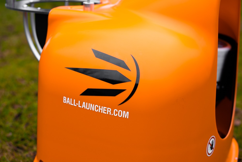 Pomarańczowa maszyna do wyrzucania piłek z logo Ball-Launcher.com na tle zielonej trawy.