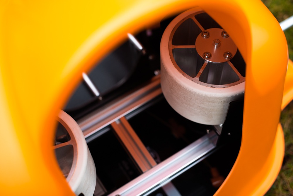 Wnętrze pomarańczowego urządzenia z dwoma białymi kołami z metalowymi elementami mocującymi, widoczne szyny i czarne elementy konstrukcyjne, oświetlone jasnym światłem.