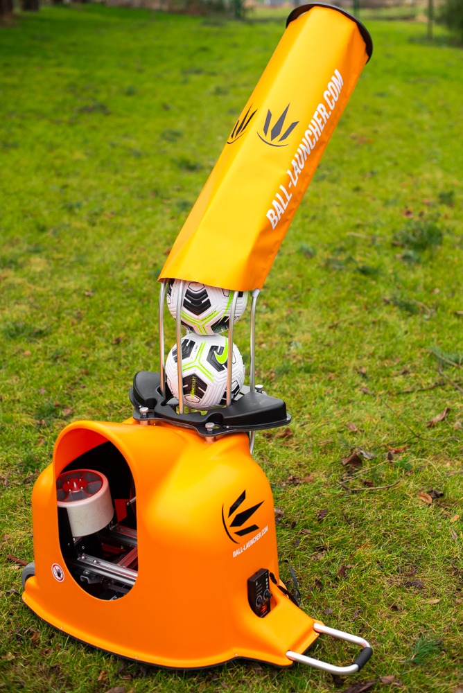 Pomarańczowa wyrzutnia piłek z dwoma piłkami nożnymi wewnątrz, stojąca na trawie, z widocznym logo i adresem strony internetowej.