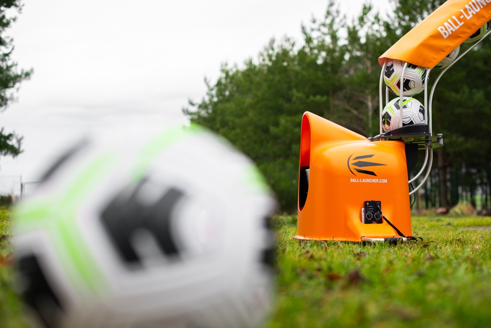 Pomarańczowa wyrzutnia piłek z trzema piłkami w podajniku, częściowo widoczna piłka w pierwszym planie, na trawie, w tle drzewa.