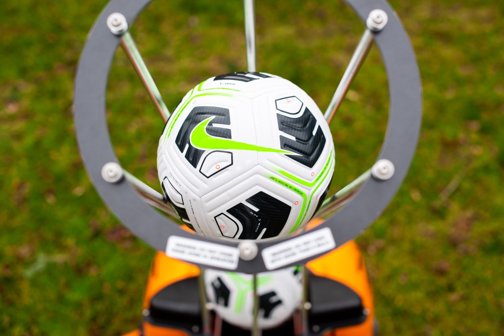 Nowa piłka nożna Nike Flight z jaskrawozielonym logo umieszczona w metalowej obręczy na tle trawy.