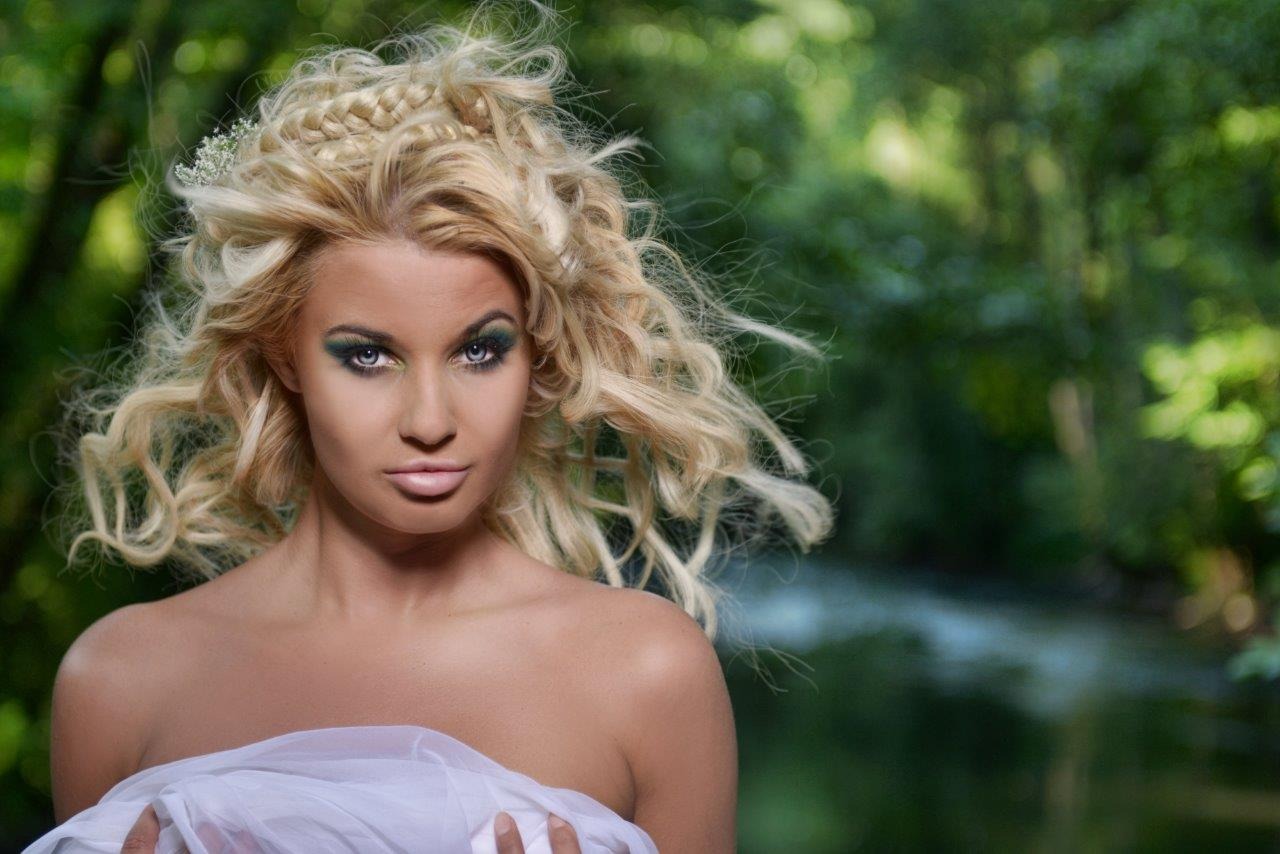 Portret kobiety z blond włosami upiętymi w warkocze i intensywnym zielonym makijażem oczu, na tle rozmytej zieleni.