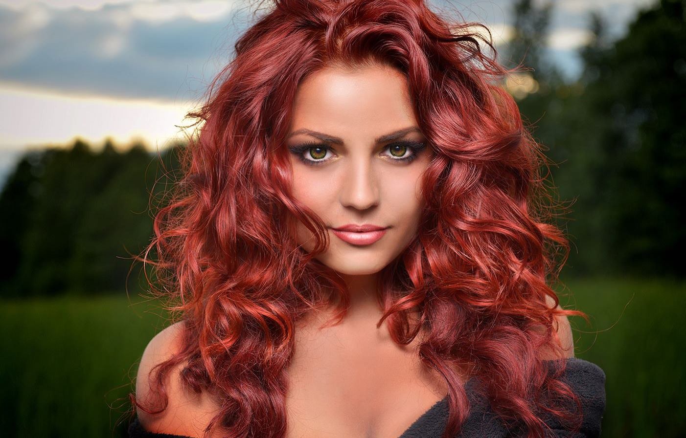 Portret kobiety z intensywnie rudymi, kręconymi włosami i wyrazistym makijażem oczu, na tle zieleni i zachmurzonego nieba.