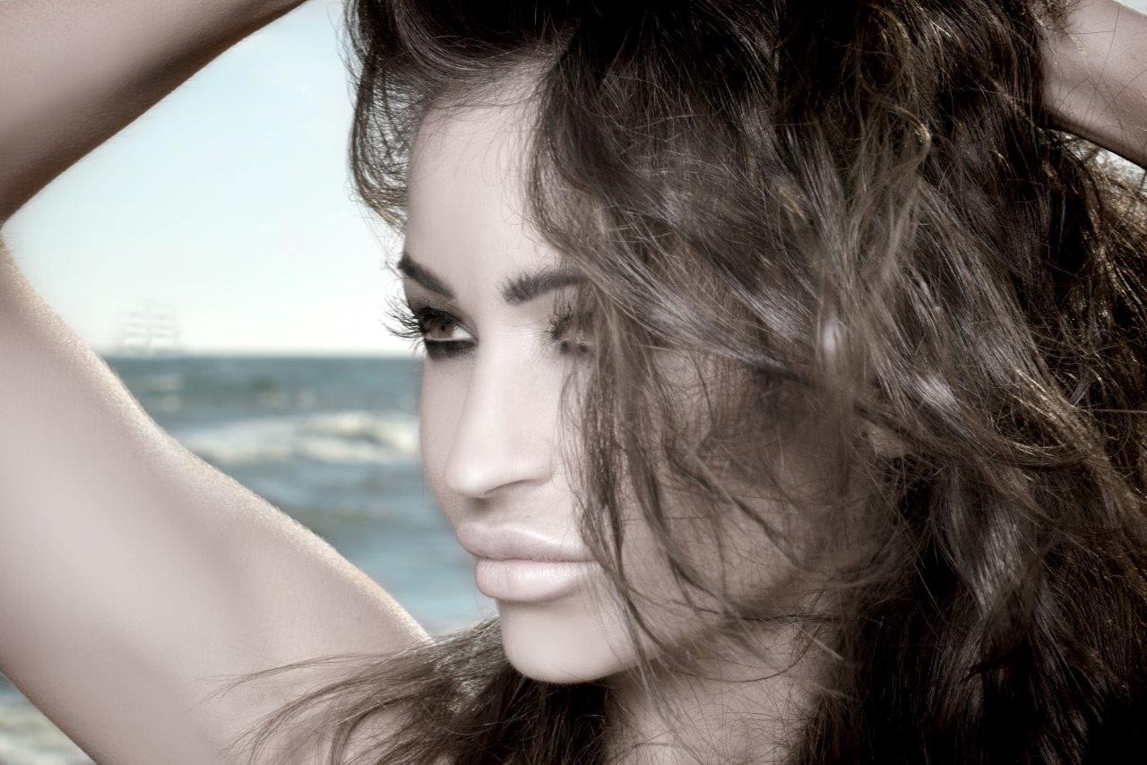 Portret kobiety z intensywnym makijażem oczu i pełnymi ustami na tle morza z żaglówką w oddali.