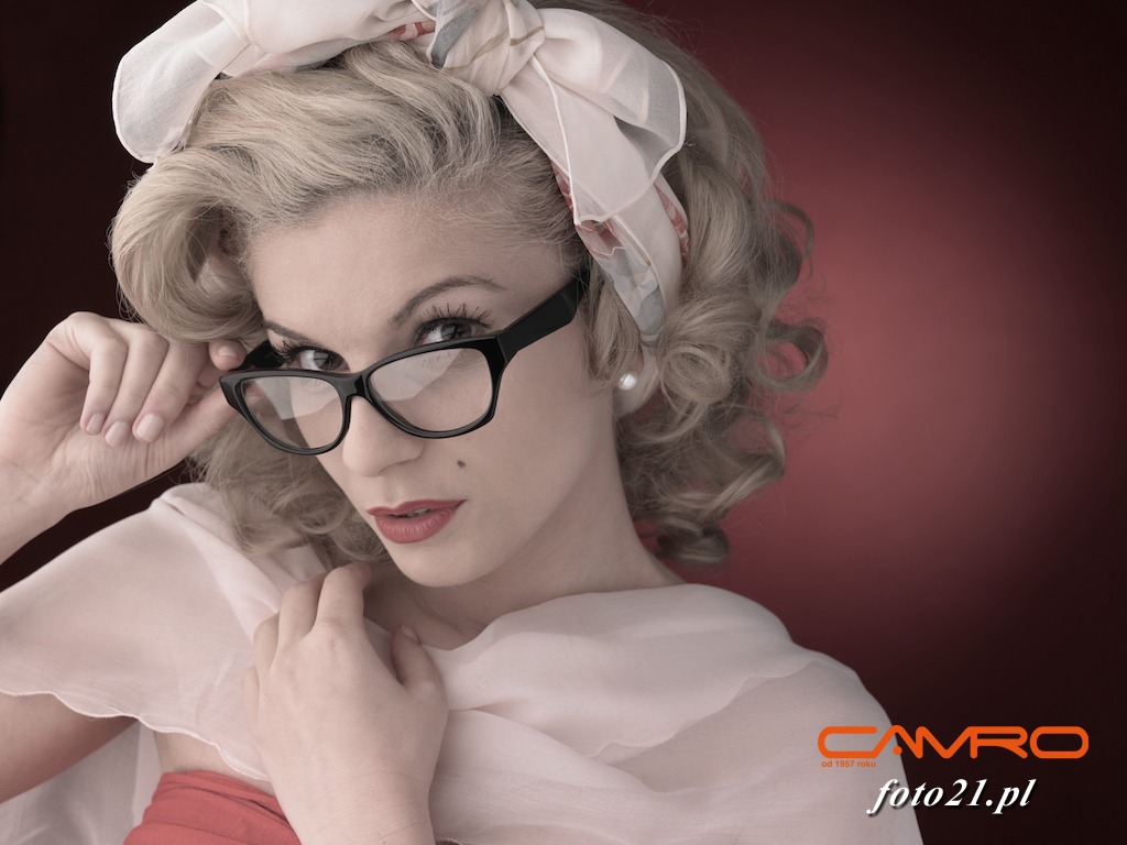 Portret kobiety w stylu retro z blond lokami, w okularach z czarną oprawką, upiętą chustą we włosach, z delikatnym makijażem i czerwonymi ustami na tle ciemnoczerwonym.