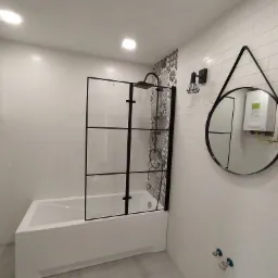 Nowoczesna łazienka z białą wanną z czarną kabiną prysznicową, okrągłe lustro z czarną ramą odbijające podgrzewacz wody, białe płytki na ścianach i designerskie lampy.