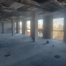 Wnętrze w trakcie remontu: surowy strop z widoczną instalacją, szpachlowane ściany, okna z widokiem na miasto, na podłodze profile i płyty karton-gips.