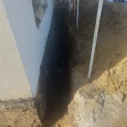 Wykop fundamentowy wzdłuż ściany budynku z widocznymi podporami i gruntem.