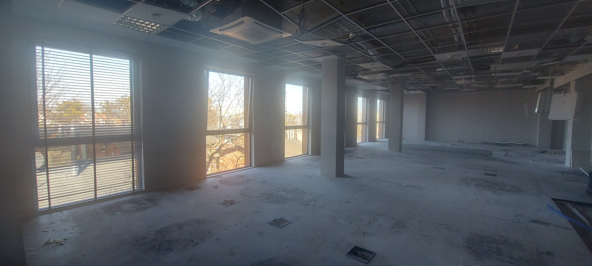 Duże, puste pomieszczenie w stanie surowym, z widocznymi instalacjami pod sufitem i kilkoma oknami z żaluzjami, przygotowane do dalszych prac wykończeniowych.
