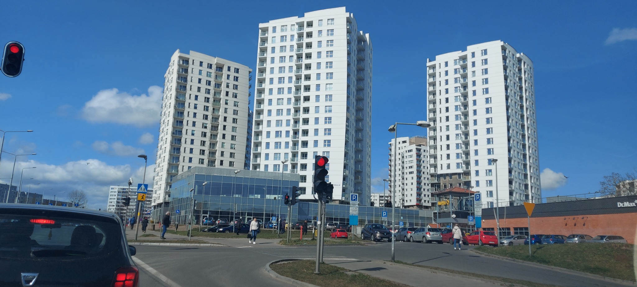 Nowoczesne, białe budynki mieszkalne z balkonami, widoczne z ulicy z sygnalizacją świetlną i zaparkowanymi samochodami w słoneczny dzień.