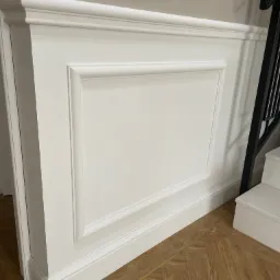 Biała sztukateria gipsowa z profilowanymi ramkami i listwami przypodłogowymi, montowana na ścianie przy schodach z parkietem ułożonym w jodełkę.