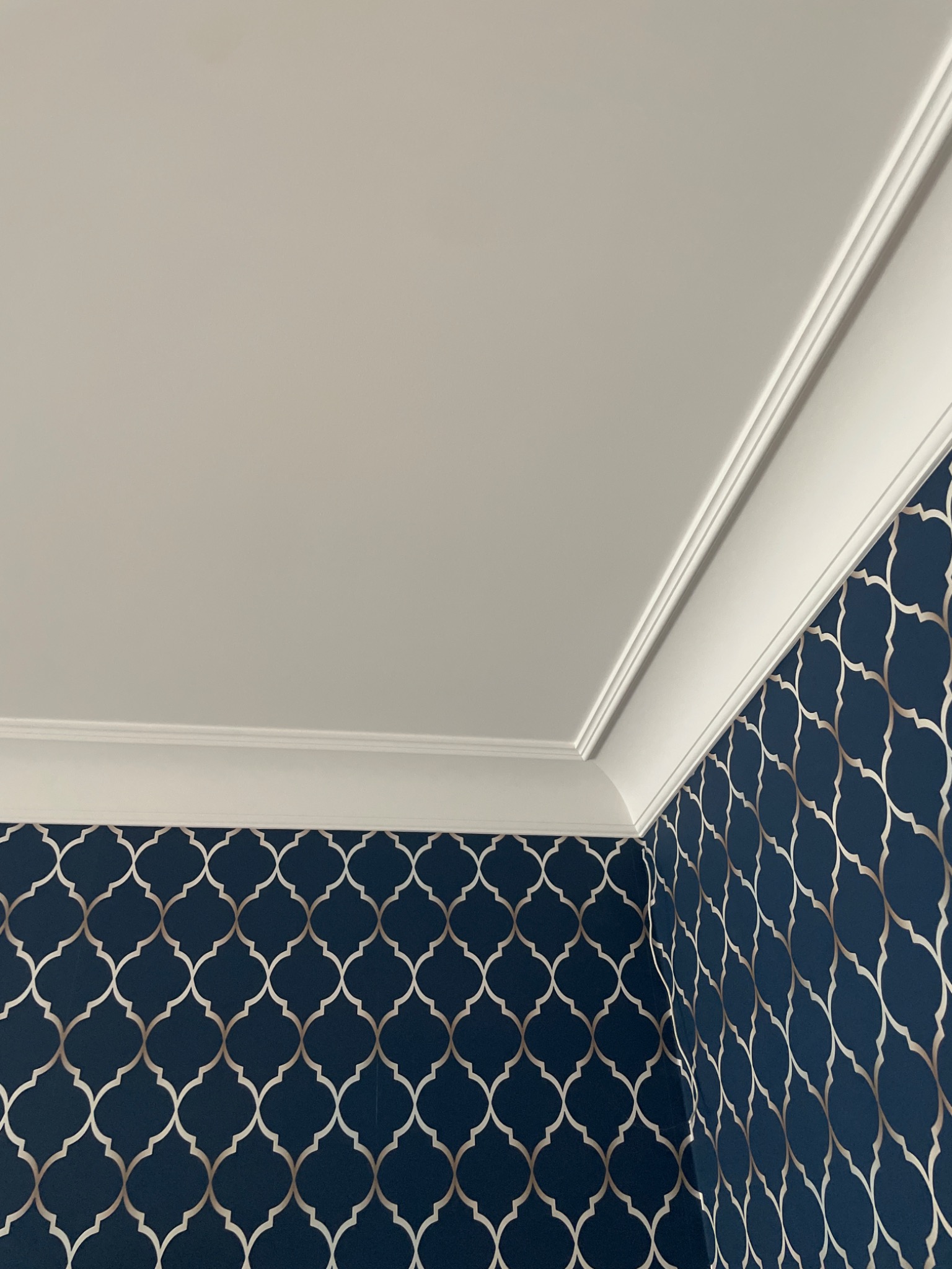 Fragment narożnika pokoju z gładkim, jasnym sufitem, dekoracyjną listwą przysufitową i ścianą pokrytą granatową tapetą w srebrzysty wzór koniczyny marokańskiej.