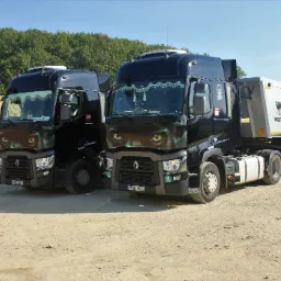 Dwa czarne samochody ciężarowe marki Renault z naczepami stoją na placu budowy, z dekoracyjną grafiką oczu na przedniej masce, w tle hałda piasku i drzewa.