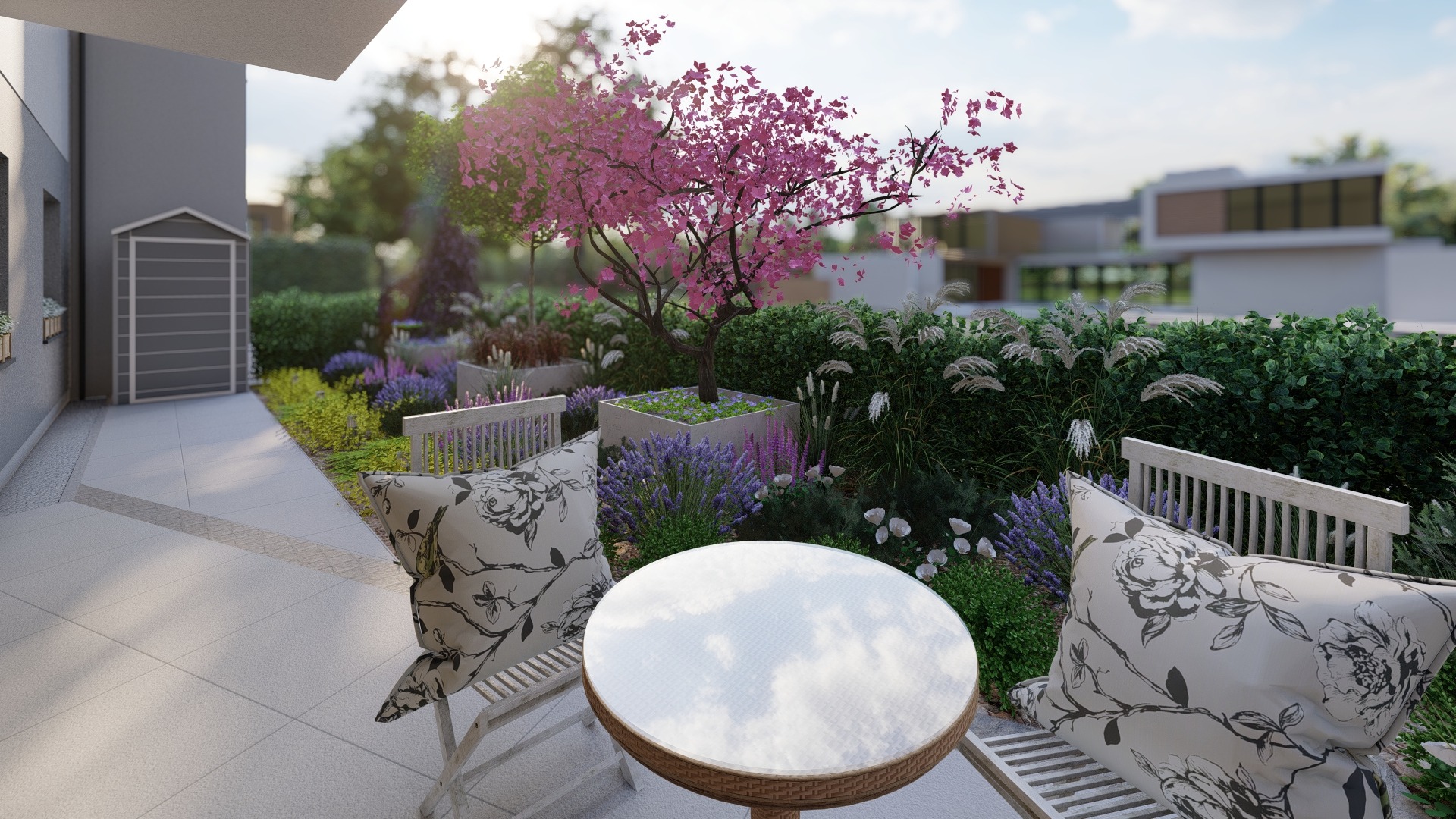 Taras z okrągłym stolikiem i krzesłami z poduszkami w kwiaty, otoczony kwitnącą lawendą, ozdobnymi trawami i drzewem o różowych kwiatach.