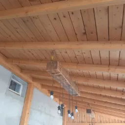 Drewniany sufit z belkami i lampą wiszącą wykonaną z belki owiniętej liną jutową, z widocznym fragmentem ściany budynku z oknem.