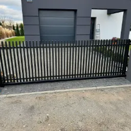 Nowoczesna, czarna brama wjazdowa na posesję z szarego bruku, na tle domu z garażem i zieleni. Minimalistyczny styl, pionowe pręty.