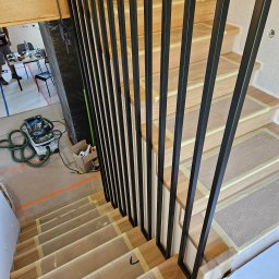 Mobilne Usługi Ślusarskie ŁUKASZ PILACZYŃSKI - Drewniane schody z czarną, metalową balustradą w trakcie montażu. Widoczne zabezpieczenia stopni i narzędzia w tle. Ujęcie z góry, w trakcie realizacji.