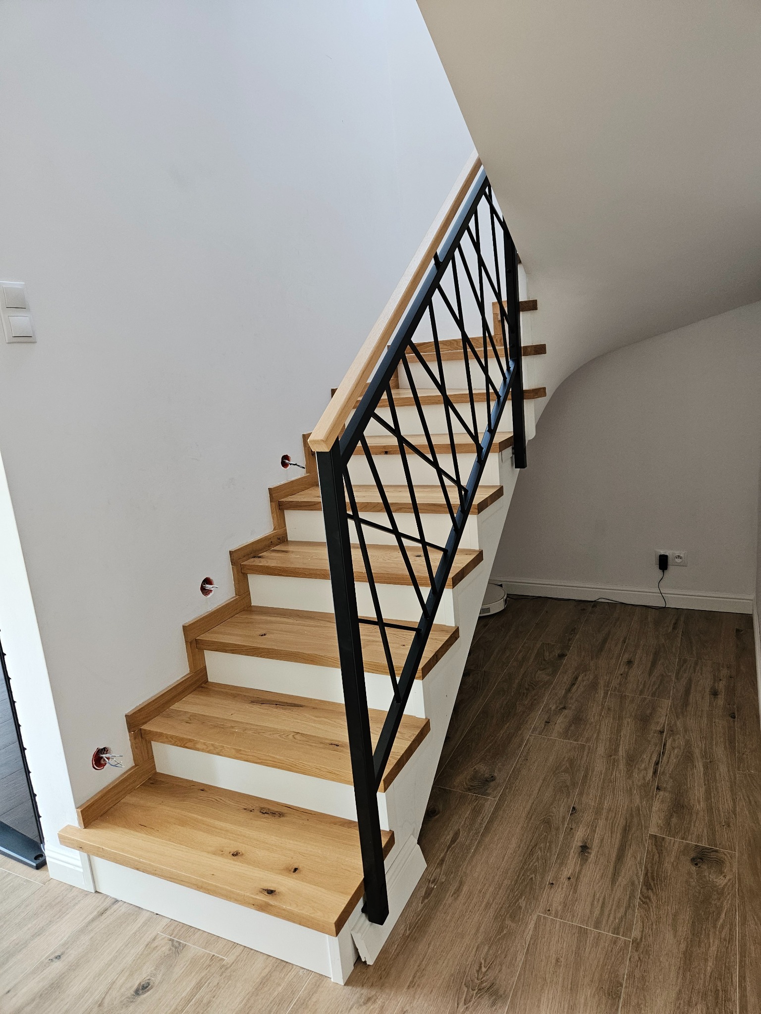Drewniane schody z czarną, metalową balustradą o geometrycznym wzorze. Jasne stopnie kontrastują z ciemną balustradą. Widoczne gniazdka elektryczne na ścianie.