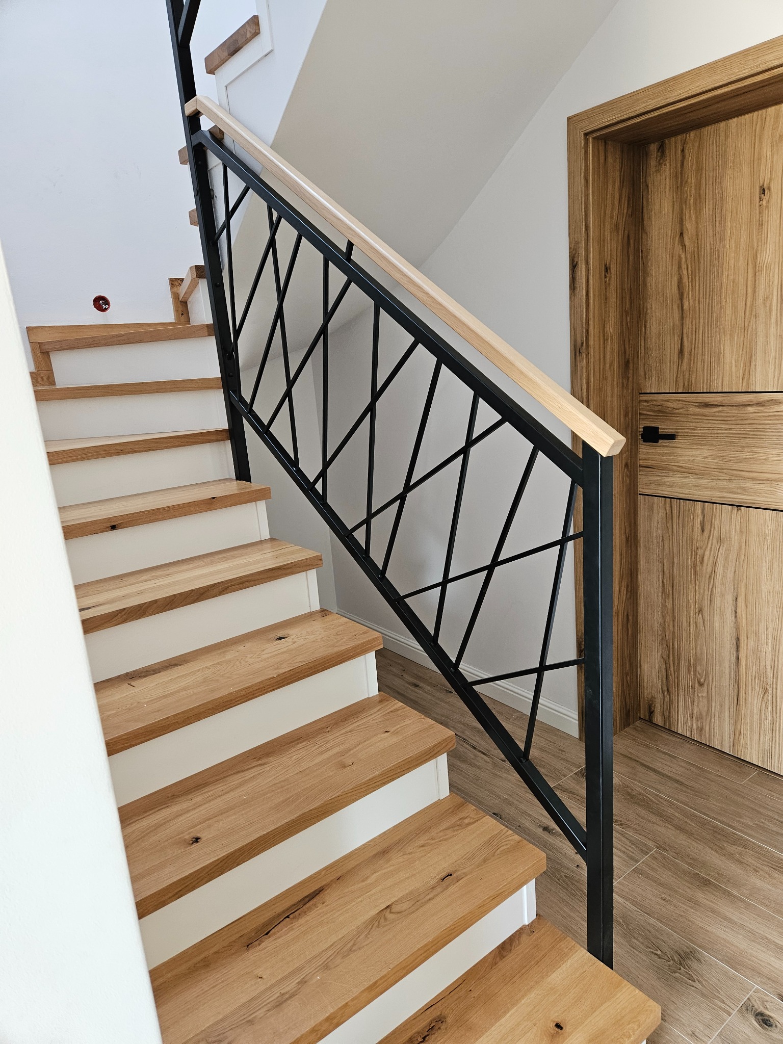 Nowoczesne schody z drewnianymi stopniami, białą podstopnicą i czarną, geometryczną balustradą. Dębowa podłoga i drzwi w tle. Jasne wnętrze.