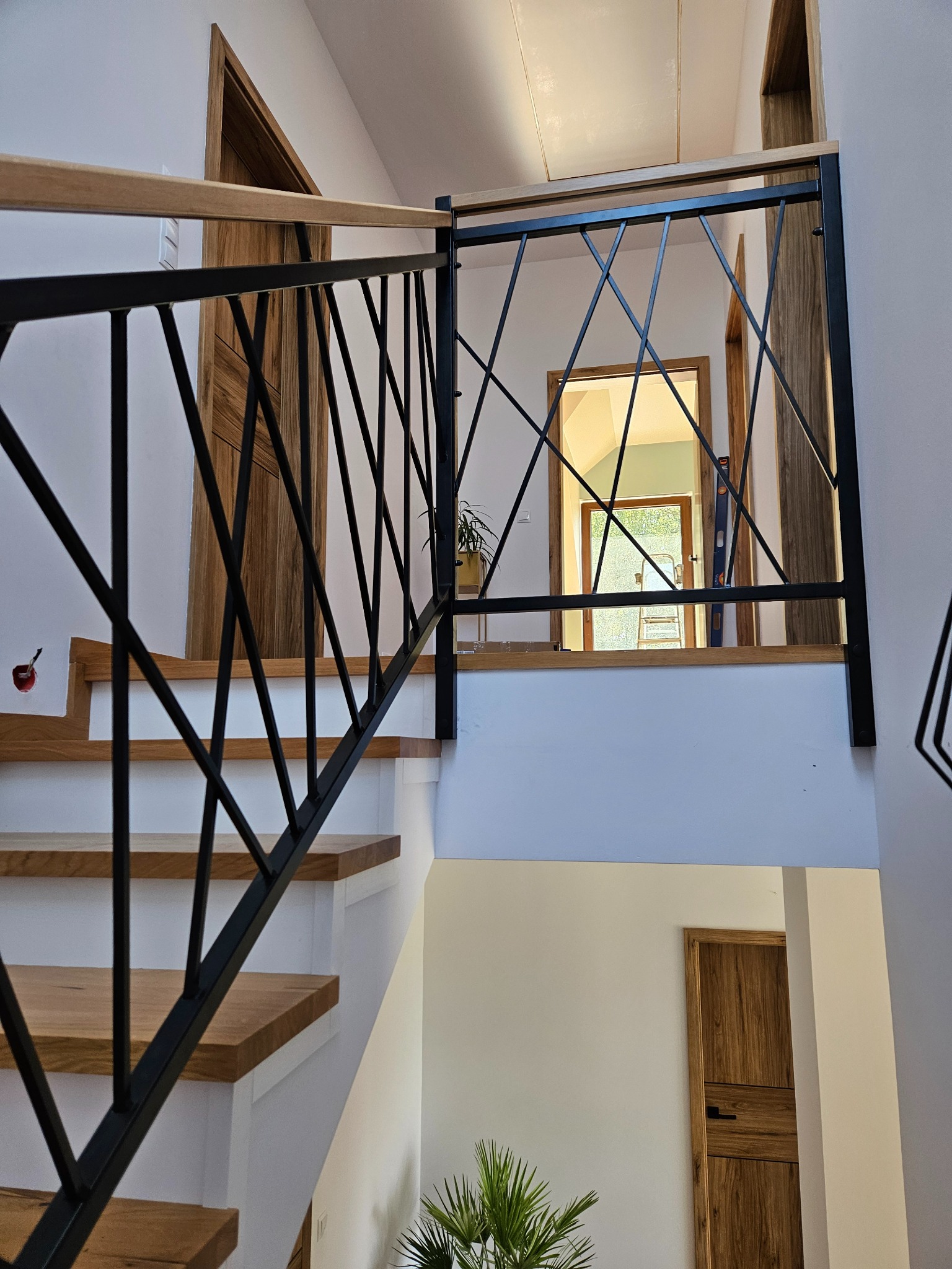 Nowoczesne schody z drewnianymi stopniami i czarną, geometryczną balustradą metalową. W tle widoczne drzwi i roślina doniczkowa.