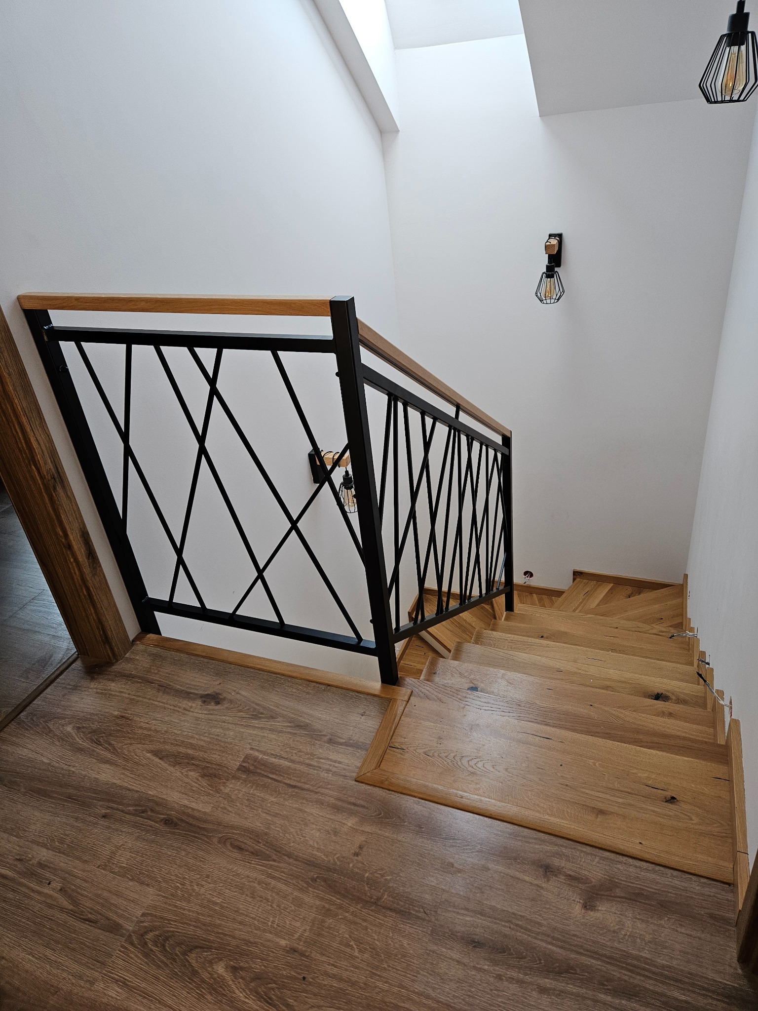 Nowoczesne schody drewniane z czarną, geometryczną balustradą metalową i drewnianym poręczą. Widok z góry, oświetlenie punktowe.