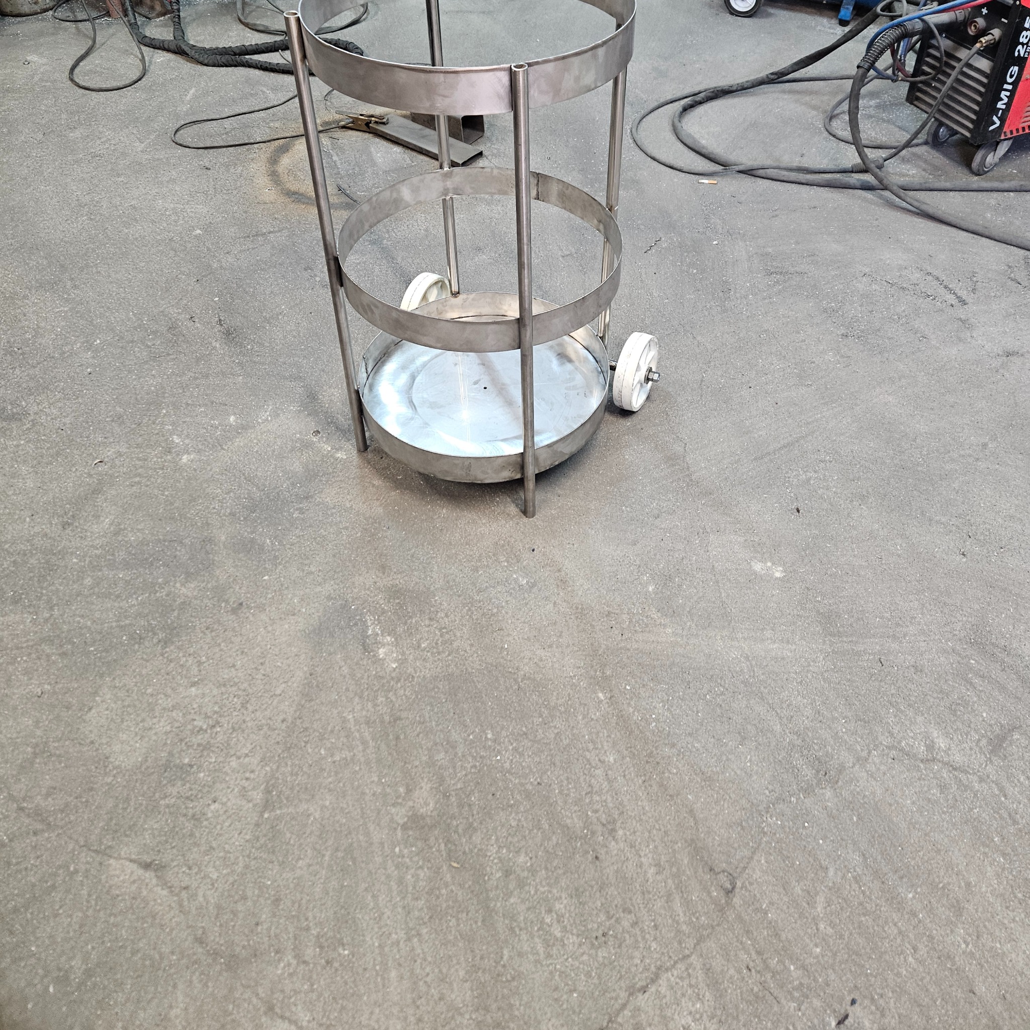 Metalowa konstrukcja na kółkach z trzema okrągłymi poziomami, spawana, surowa, w warsztacie. Widoczny spawarka i przewody.