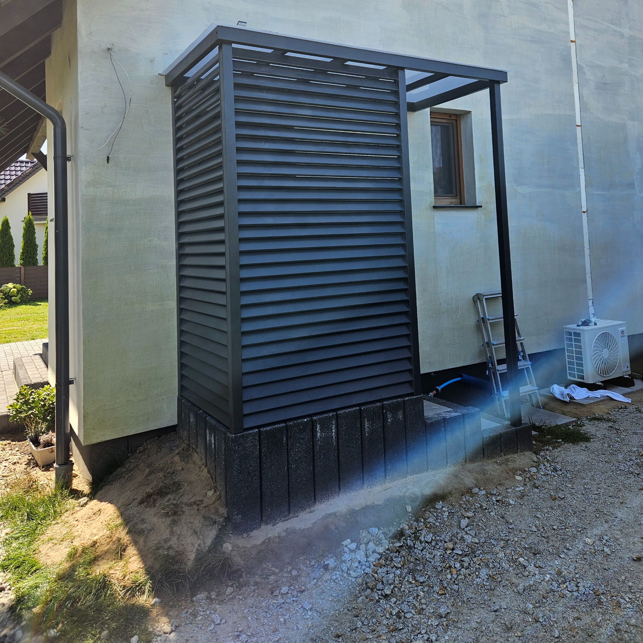 Nowoczesna, grafitowa pergola metalowa z żaluzjowymi panelami, osłaniająca element na zewnątrz budynku. Widoczny fragment okna i drabina oparta o ścianę.