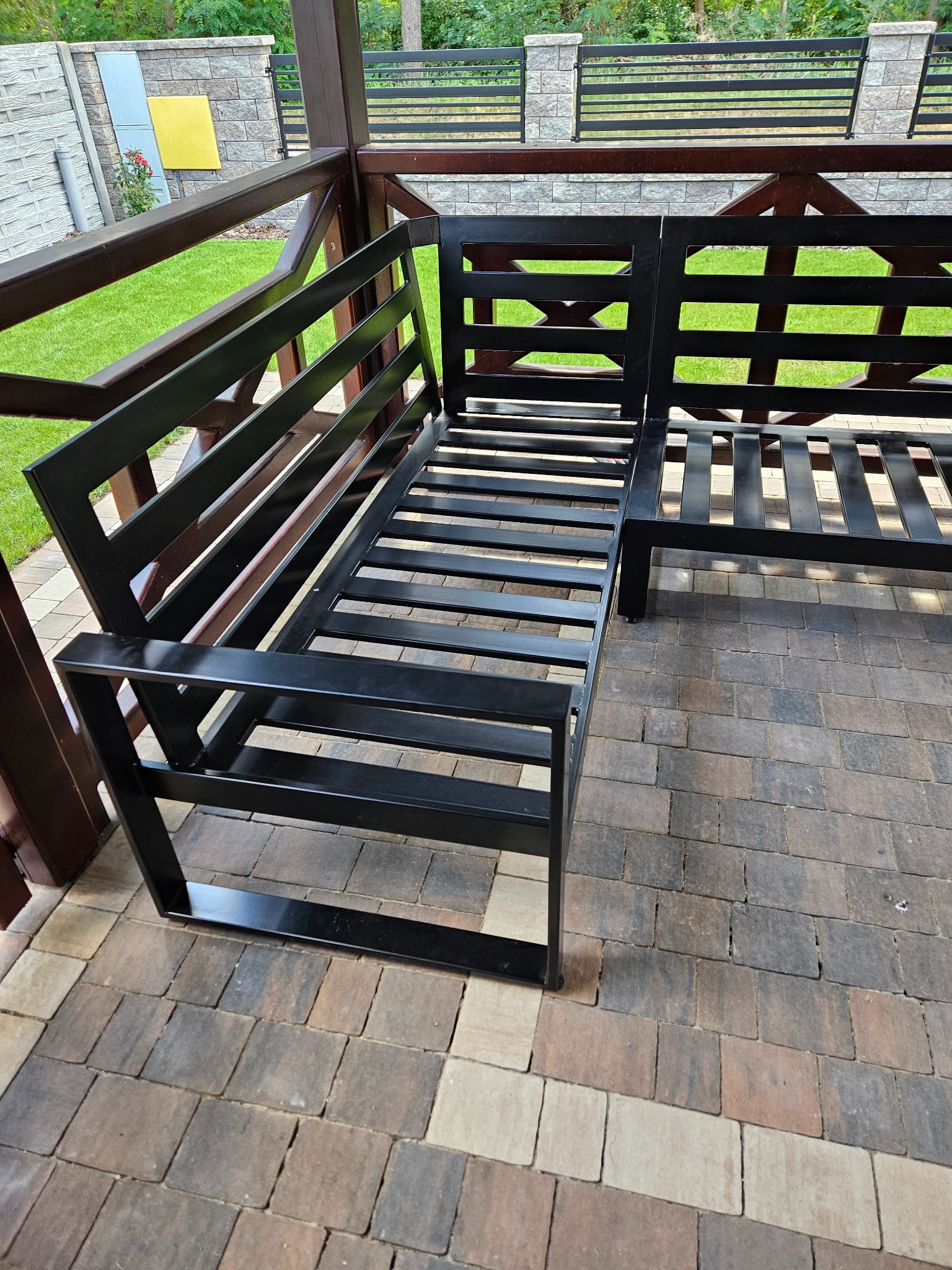 Czarna metalowa sofa na zewnątrz, z geometrycznym wzorem, umieszczona na brukowanym patio pod drewnianą pergolą. Widok na trawnik i ogrodzenie w tle.