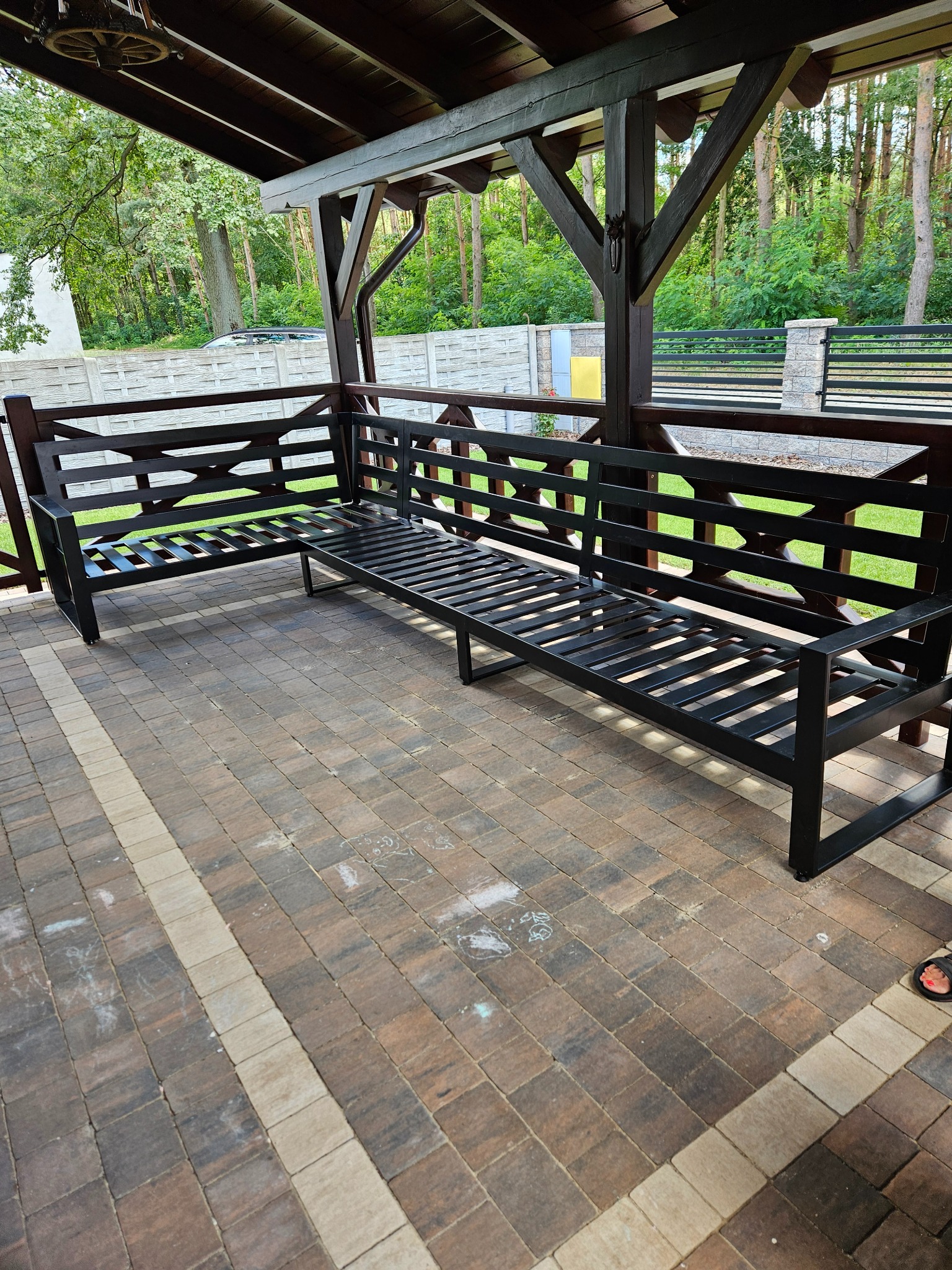 Metalowa, czarna ławka narożna na zadaszonym tarasie z kostki brukowej. Widok na zieleń za ogrodzeniem. Elementy drewniane konstrukcji zadaszenia.