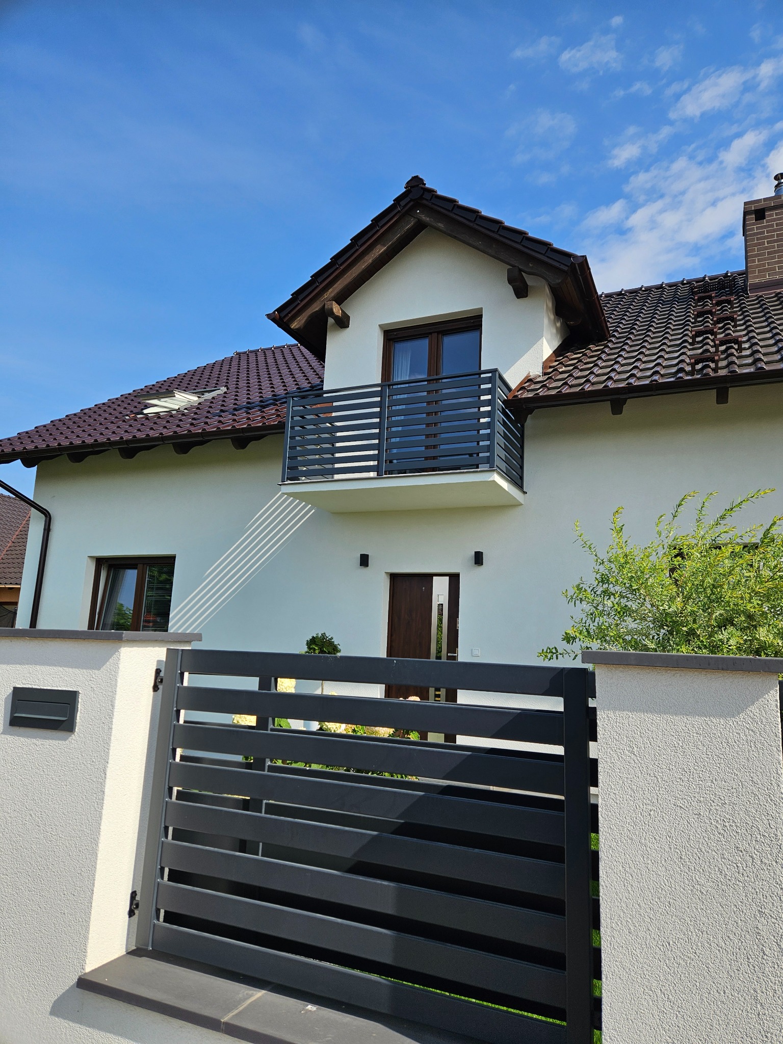 Nowoczesna, pozioma brama wjazdowa z metalu w kolorze antracytowym, na tle białego domu z balkonem i brązową dachówką, w słoneczny dzień.