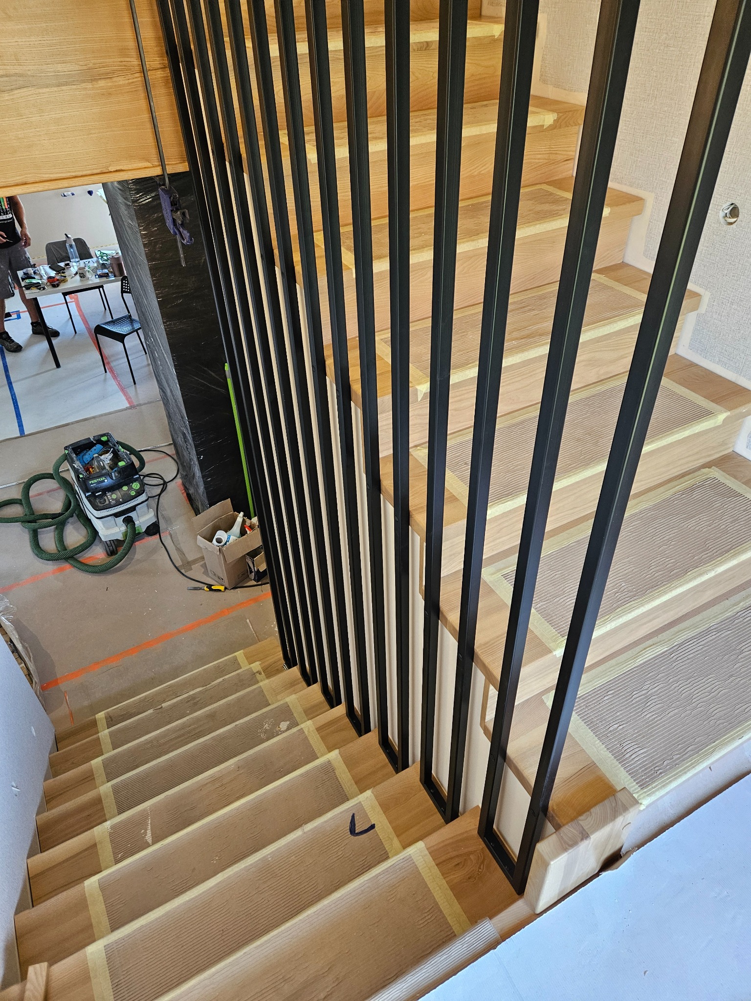 Drewniane schody z czarną, metalową balustradą w trakcie montażu. Widoczne zabezpieczenia stopni i narzędzia w tle. Ujęcie z góry, w trakcie realizacji.