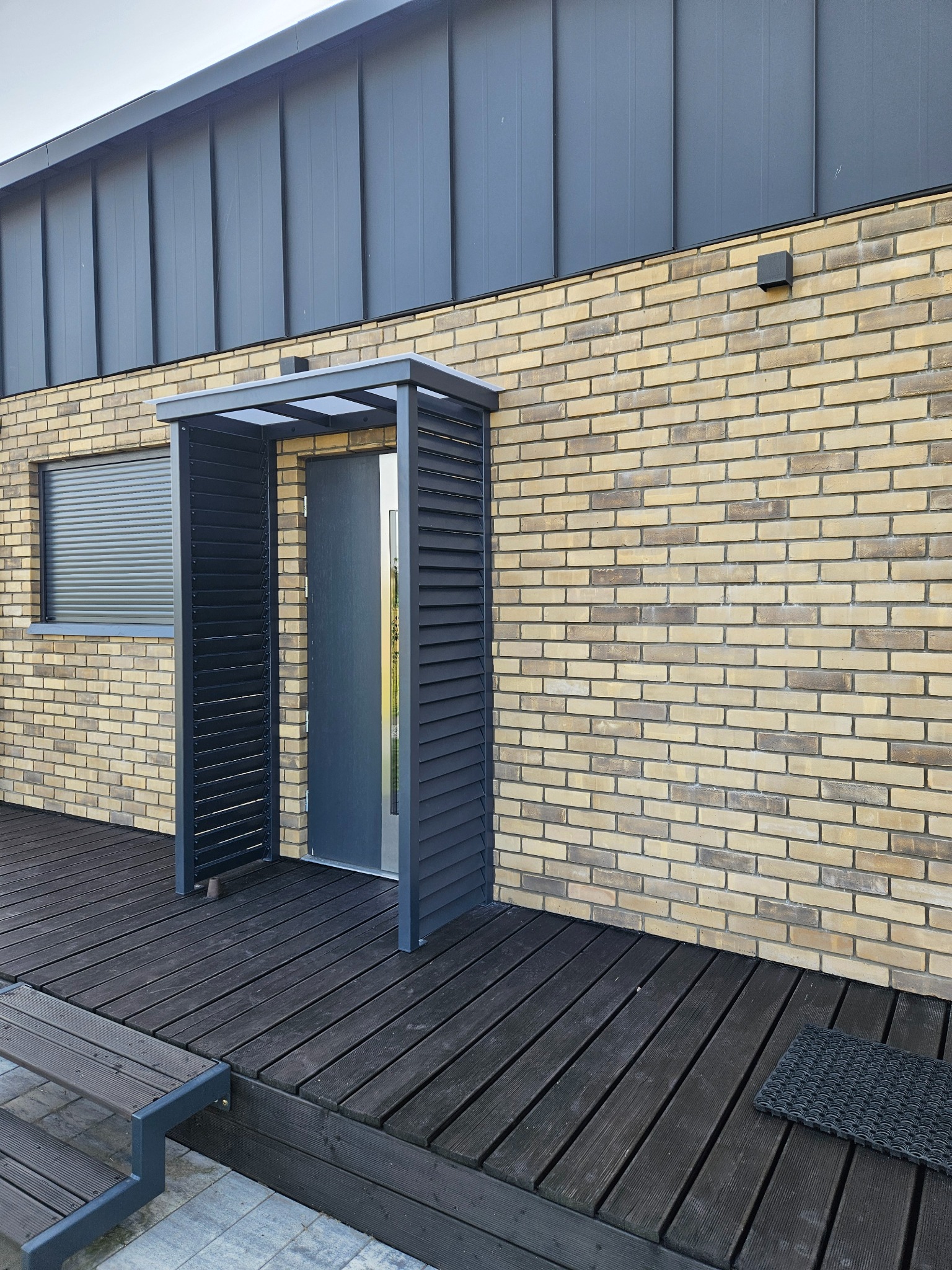 Nowoczesne zadaszenie nad wejściem do domu, z lamelami po bokach, tworzące minimalistyczną konstrukcję. Elewacja z cegły, drewniany taras i szare detale.