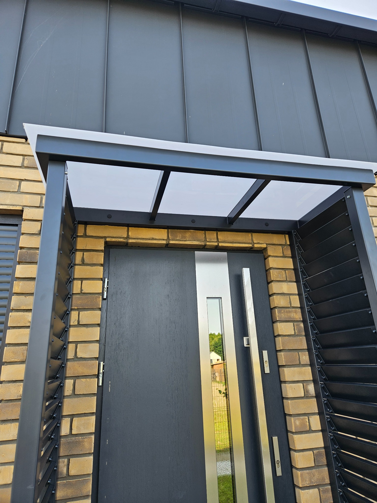 Nowoczesne zadaszenie nad drzwiami wejściowymi, wykonane z grafitowej konstrukcji metalowej i przezroczystego tworzywa, wkomponowane w elewację z cegły.